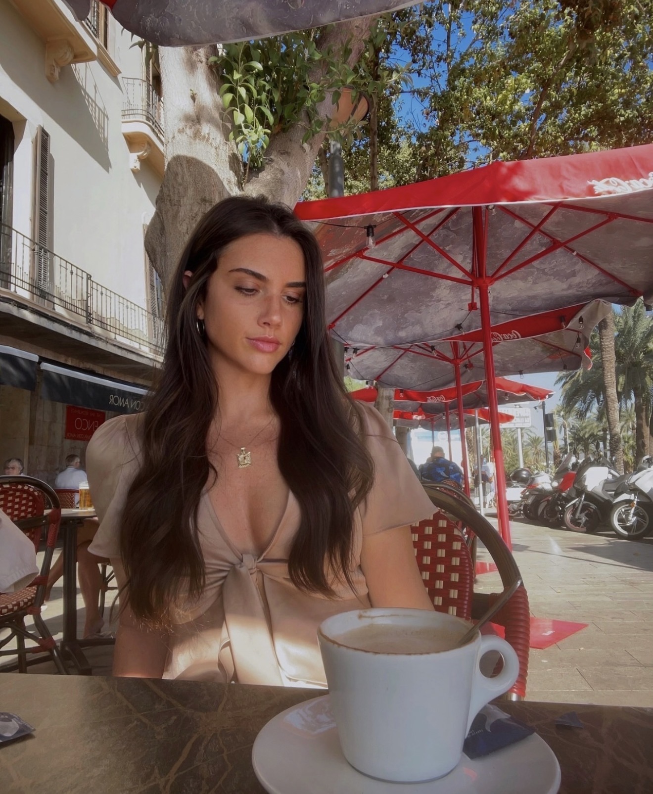 Lucia at a seaside cafe table with a cappuccino under red umbrellas