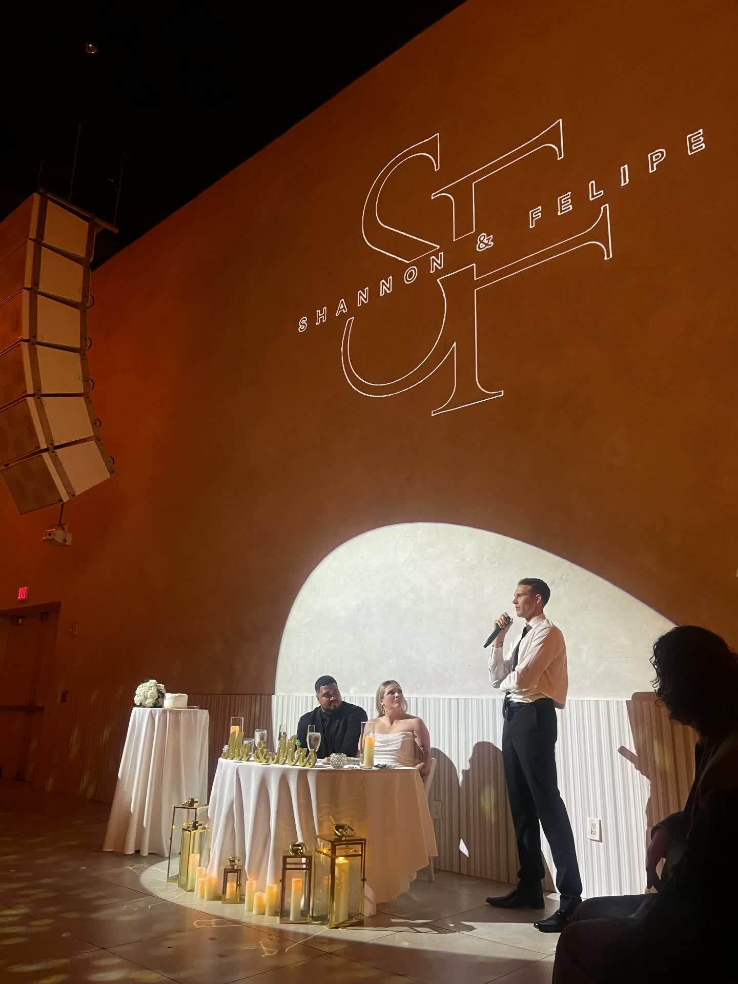 Best man giving a speech beside the couple's sweetheart table under a projected monogram