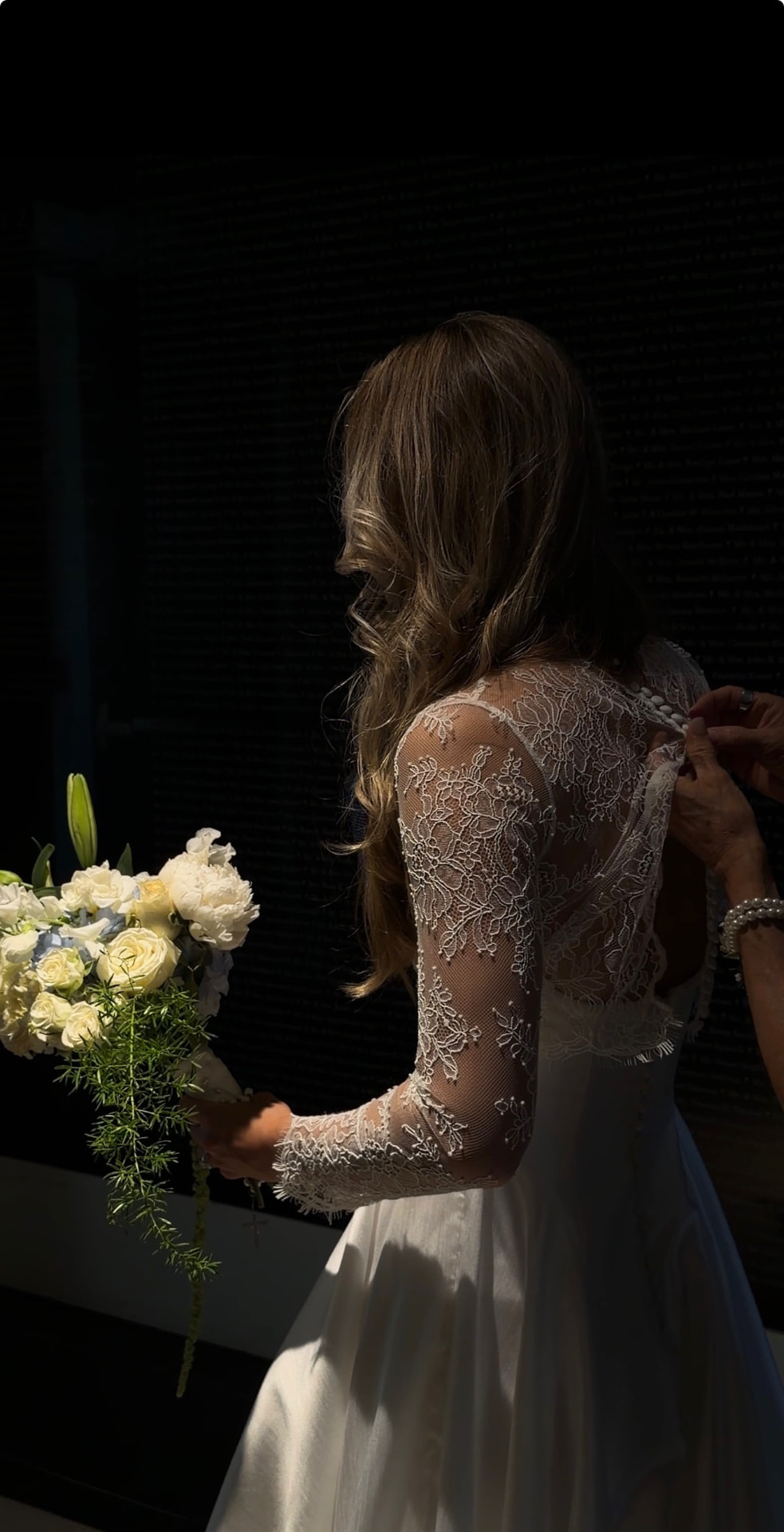 Bride in a lace-sleeved gown being buttoned in while holding her bouquet