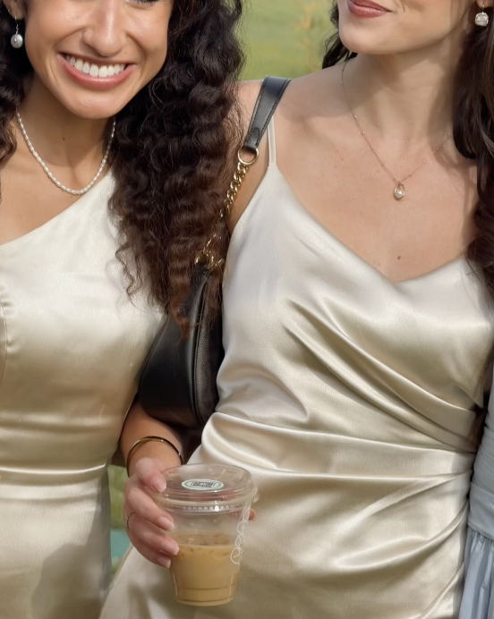 Two guests in champagne satin dresses sharing an iced coffee outdoors